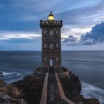 Farol iluminando o horizonte em meio a um céu dramático, representando como a fé guia e ilumina nosso caminho na vida