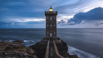 Farol iluminando o horizonte em meio a um céu dramático, representando como a fé guia e ilumina nosso caminho na vida